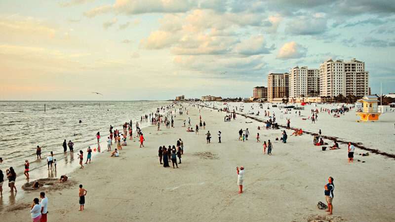 Beachgoers swim and hang out along a broad coastline with buildings in the background