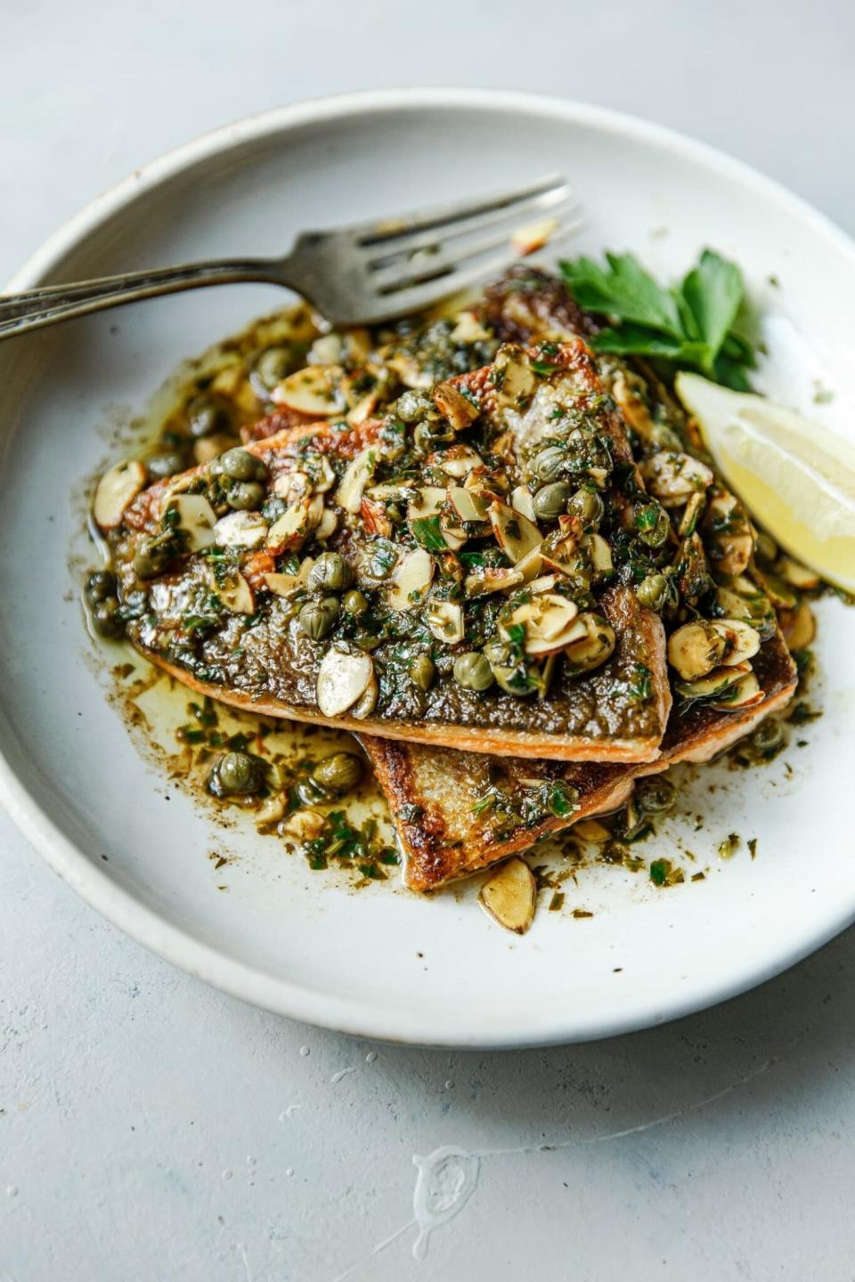 Grilled fish fillet topped with capers, sliced almonds, and herbs, garnished with lemon and parsley on a white plate with a fork