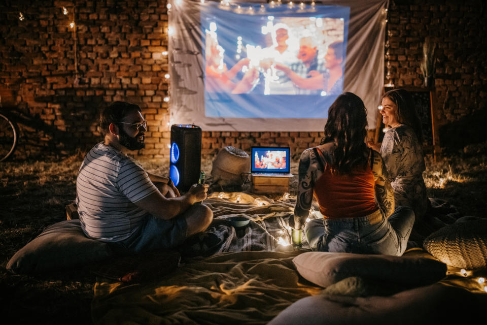 Three people, man and two women sitting together on the ground in back yard outdoors. They are watching a movie together.