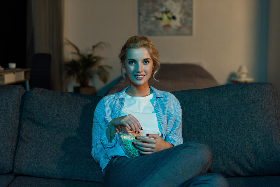 A young woman sitting on a couch watching television and eating popcorn.
