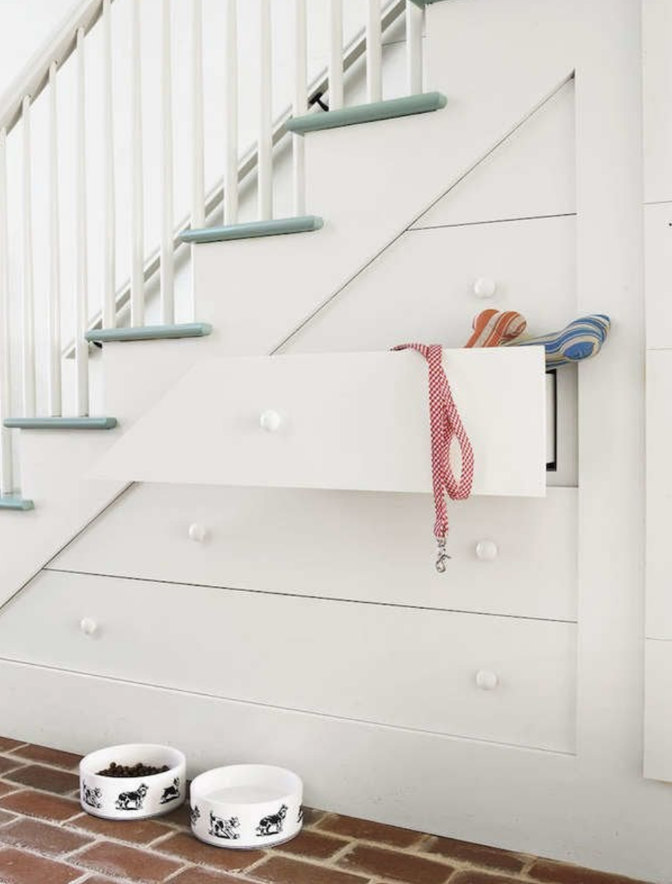 white nook under staircase with green stairs with drawers containing dog leash and toys with dog food and water bowls on the floor