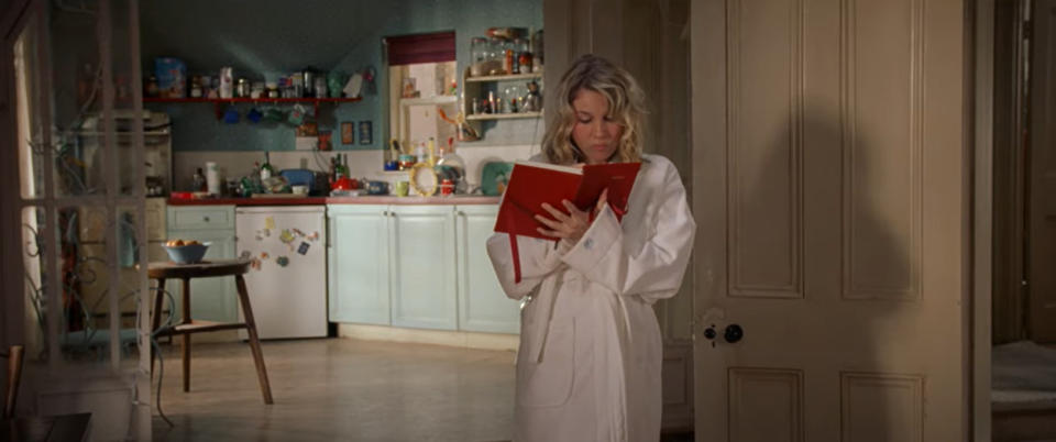 Renée Zellweger reads a red book in a kitchen wearing a white bathrobe, a scene from "Bridget Jones's Diary"