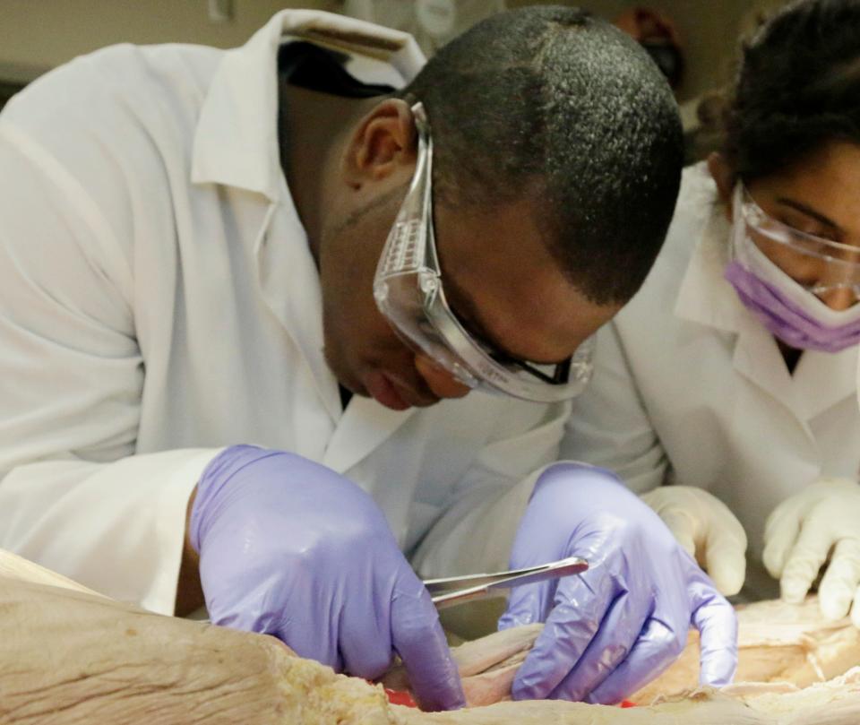 A man in a white lab coat, safety goggles, and purple medical gloves dissects a tissue sample from a cadaver.