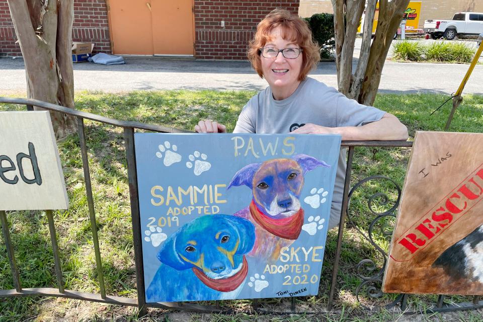 Candace Powell, manager of the Junkyard Dog thrift store in Cinco Bayou, poses with a painting of the two dogs she adopted from the Panhandle Animal Welfare Society in 2020. The painting is part of an outdoor exhibit at Junkyard Dog that features some of the animals adopted from PAWS.