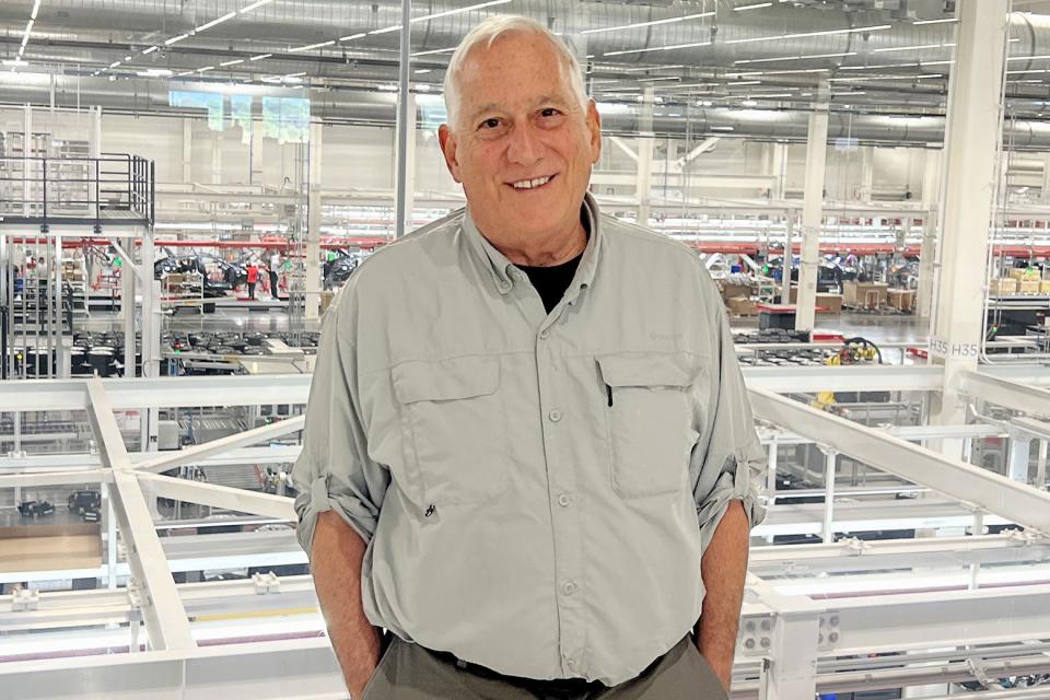 A white-haired white man wears a beige shirt and stands in a warehouse.