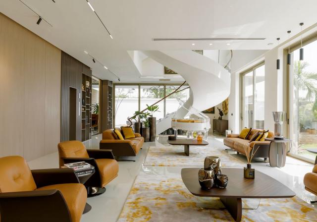 long living area with supple caramel colored leather swivel armchairs and pedestal tables and area rugs in light yellow and white and a white ceiling and glass doors and a swirly white spiral staircase at the far end
