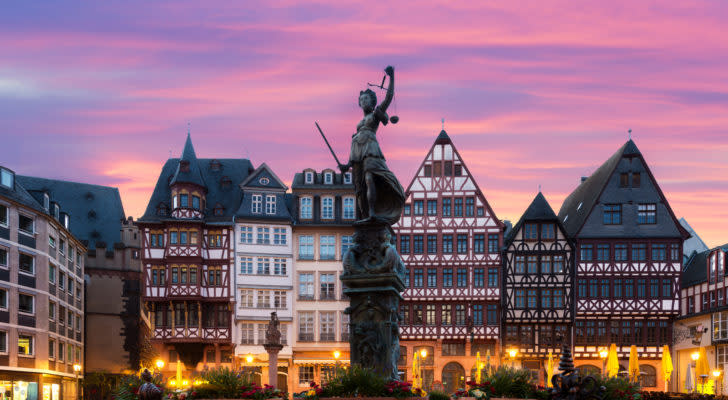 The statue of Lady Justice in Frankfurt, Germany is centered with buildings lit up behind in dusk.