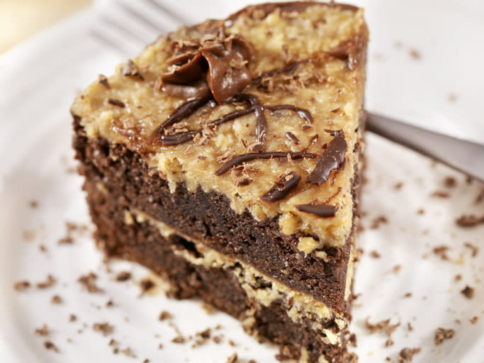 A slice of German chocolate cake with coconut-pecan frosting on a white plate, topped with chocolate drizzle and a chocolate swirl. A fork rests beside it