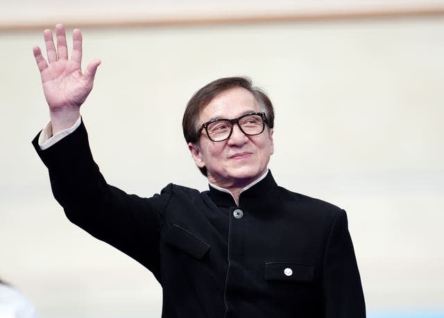 Jackie Chan, in a black jacket, waves