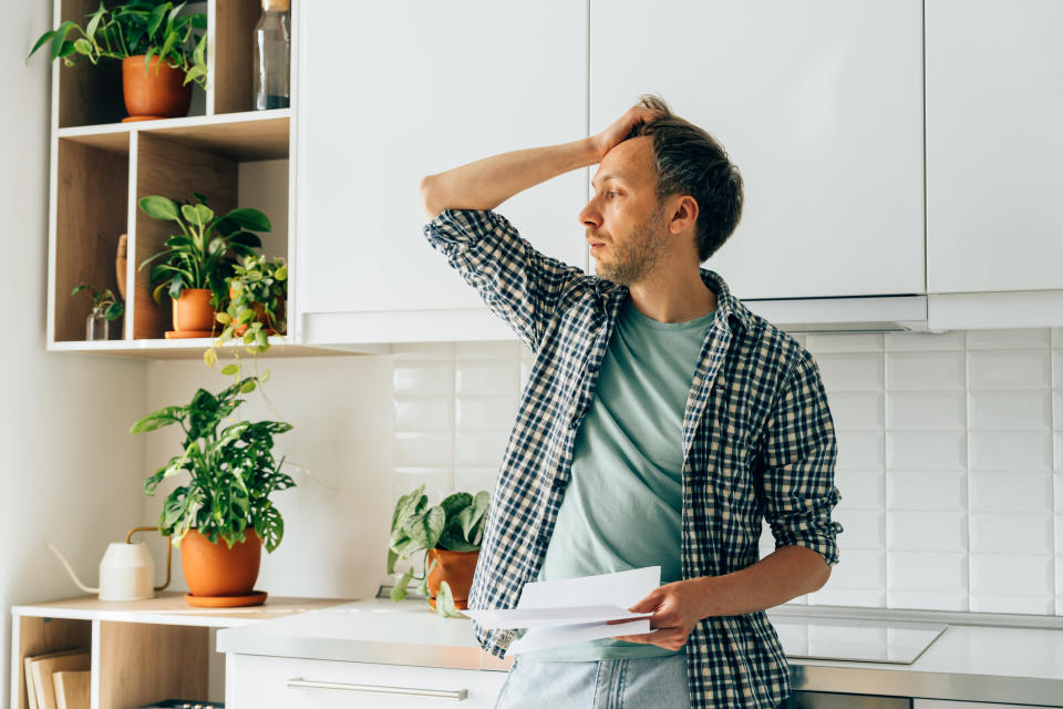 Man in a plaid shirt standing in a kitchen, looking thoughtful with paper in hand