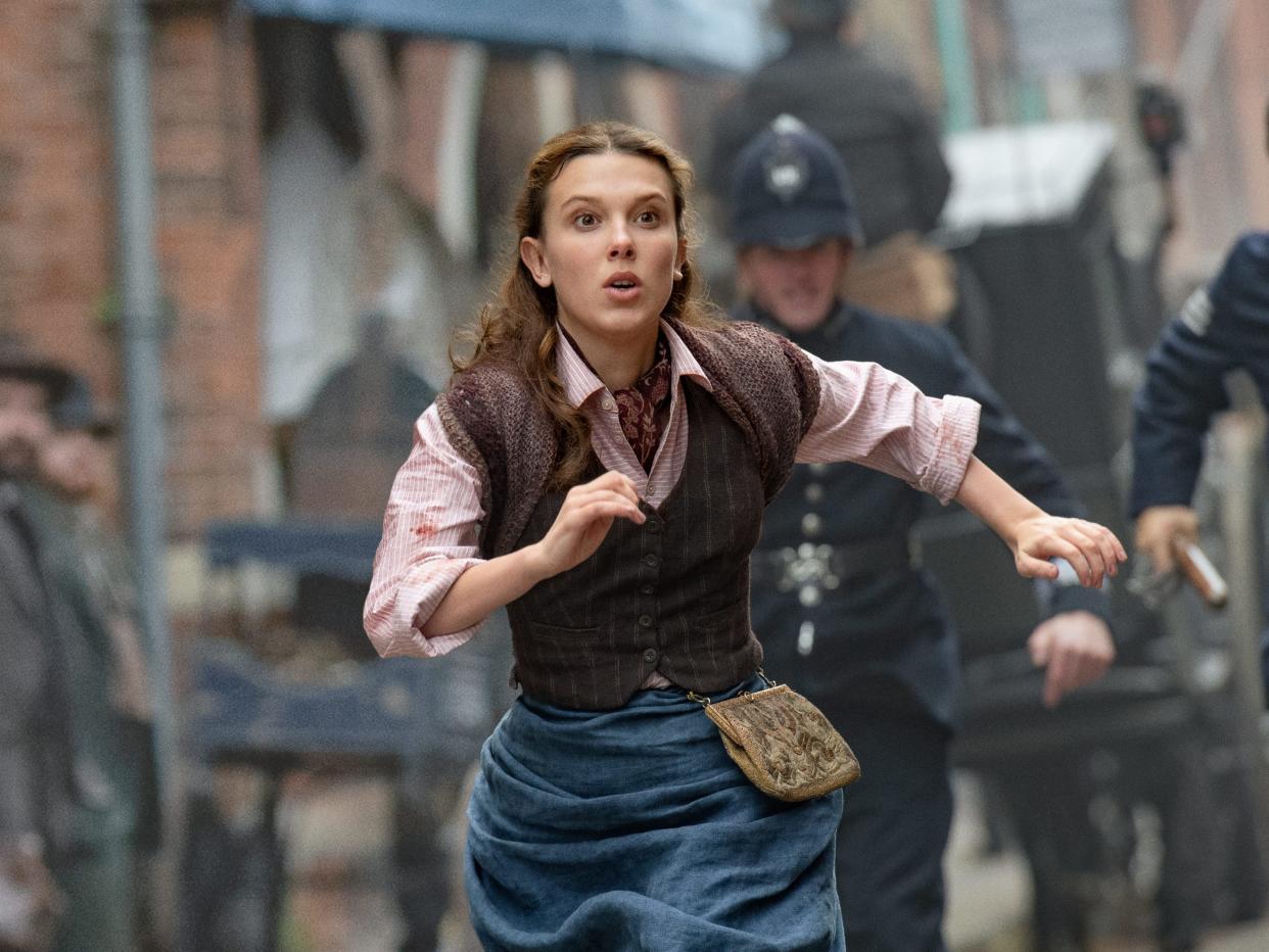 millie bobby brown as enola holmes in enola holmes two, running through the cobblstone streets of victorian london in working class clothes, police hot on her heels