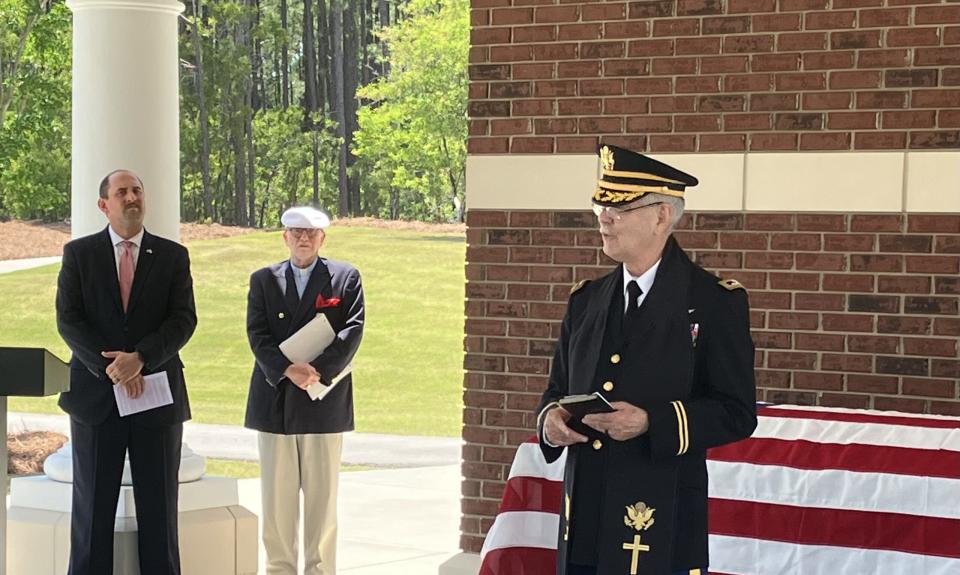 A man in a black military uniform and chaplains stole stands in front of a flag-draped coffin. Two men in suits stand off to the left side.