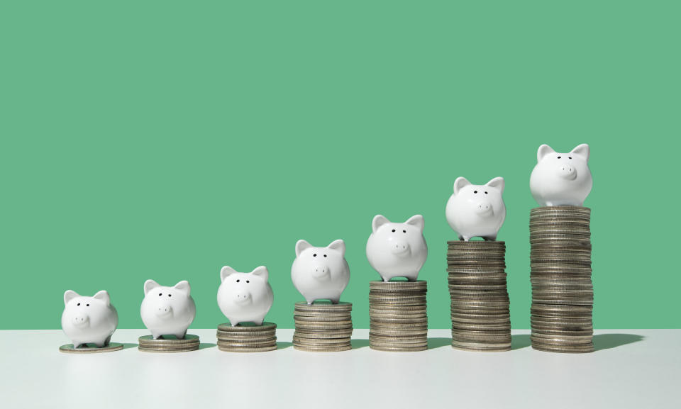 Little white piggy banks standing on top of 7 stacks of coins in ascending order on white surface, green background