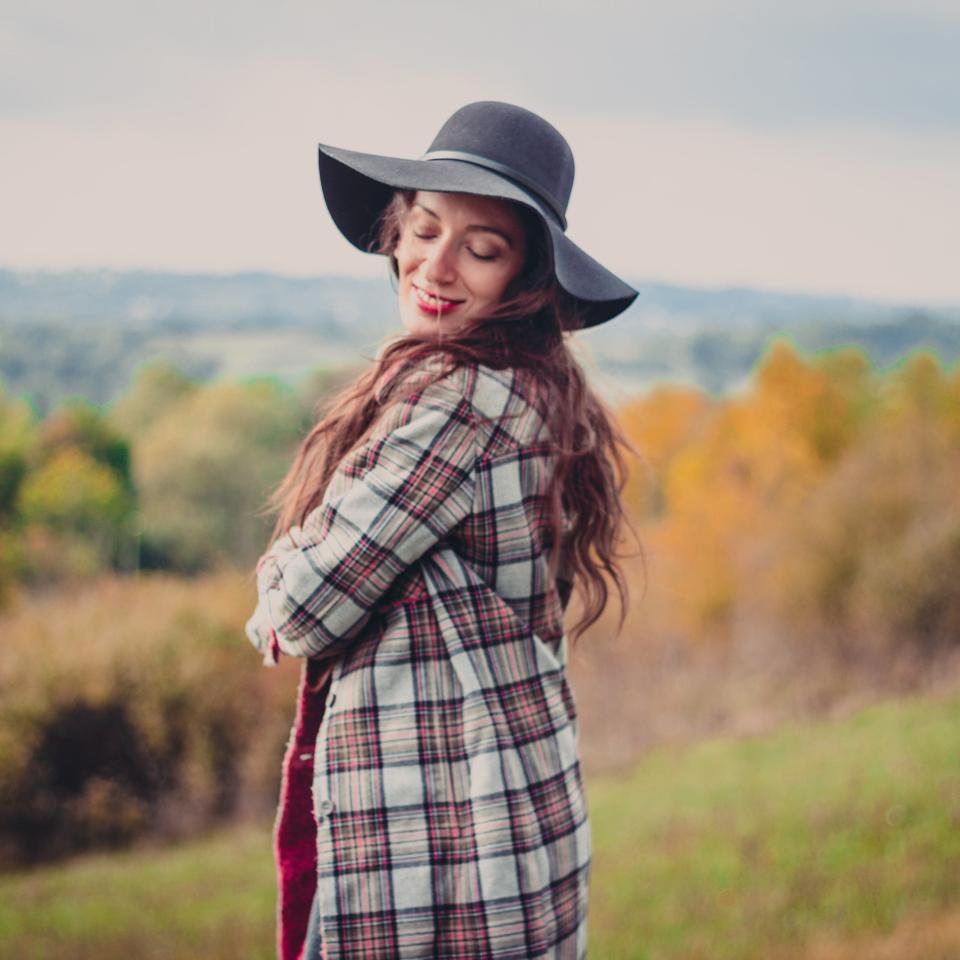 Woman in a checkered coat and wide-brim hat stands in a grassy field with autumn foliage in the background. She smiles and closes her eyes. Names unknown