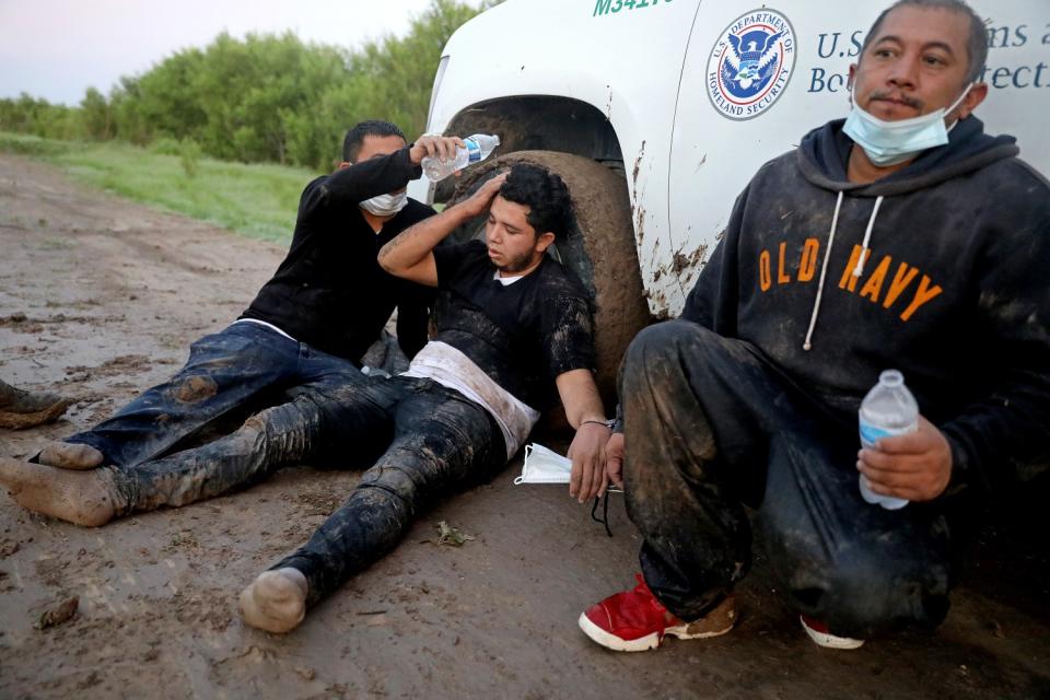 A man pours a bottle of water over another man's head as they sit on a muddy ground next to a third man