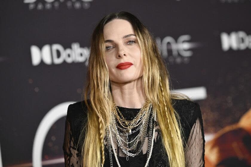 A woman with long blond hair wearing a dark dress and multiple necklaces posing against a dark background