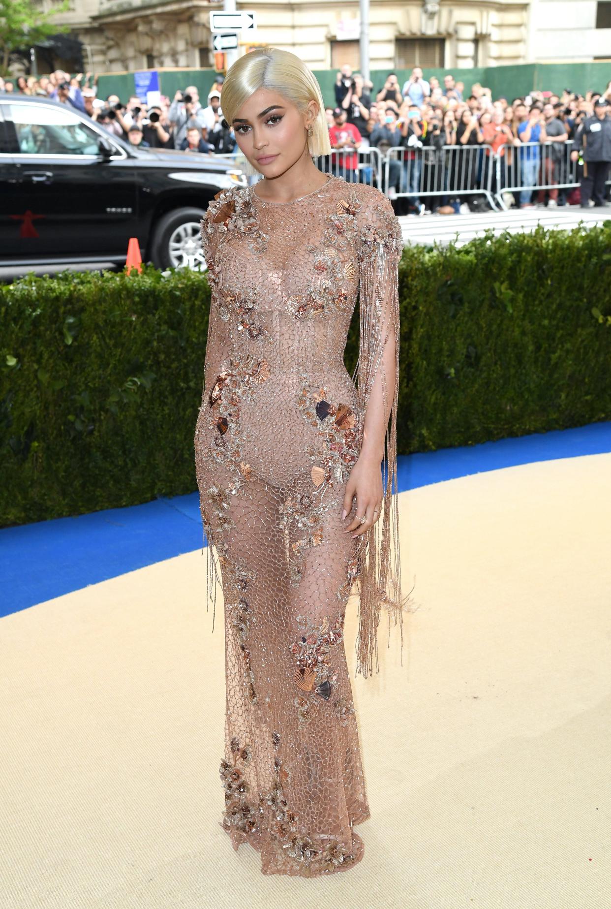 Kylie Jenner poses on the Met Gala carpet wearing a sheer, nude ensemble embellished with crystal and copper accents. Her hair is a blonde bob.