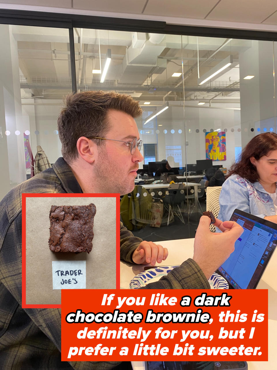 Man in glasses and plaid jacket sitting at a table with his computer, holding a piece of a brownie