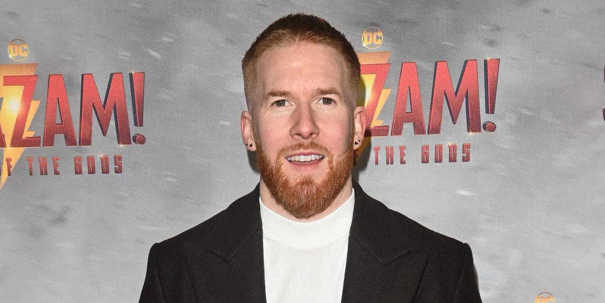 neil jones, a man stands with his hands behind his back looking at the camera with a slight smile, he has short ginger hair and beard, he wears a white jumper with black trousers and coat