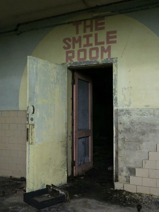 An old, partially open door with "The Smile Room" written above it on a deteriorated wall. The room inside is dark and uninviting