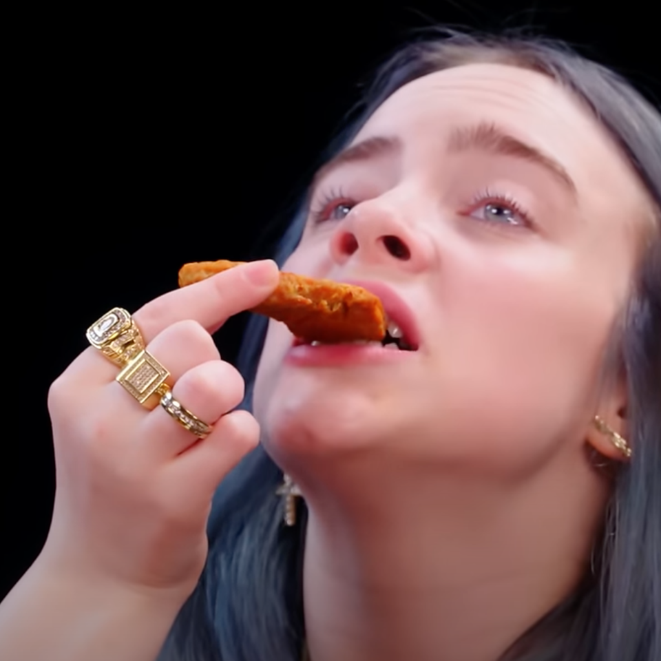 Billie Eilish eating a spicy wing, looking upwards, wearing gold rings and earrings