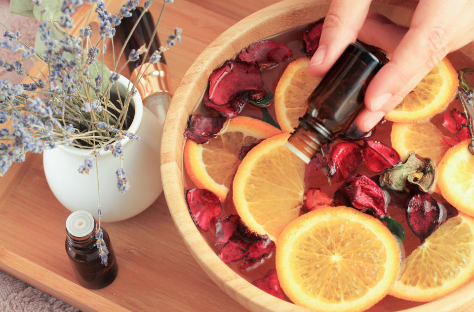 Bowl of sliced oranges and rose petals in water with a hand adding drops of essential oils to it from an apothecary bottle. Vase of dried lavender sits on a tray next to the bowl.