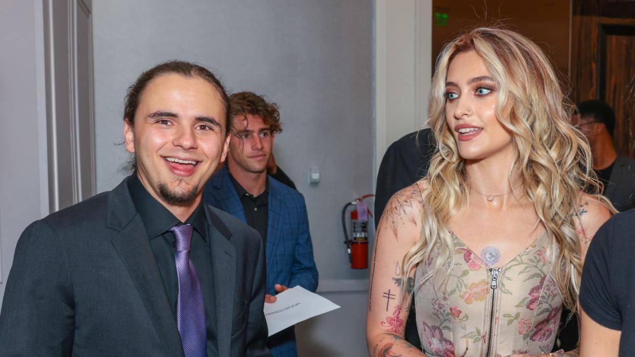 prince jackson smiles at the camera with his mouth open, he wears a gray suit jacket, dark gray collared shirt and purple tie, his hair is pulled back and another man stands in the background