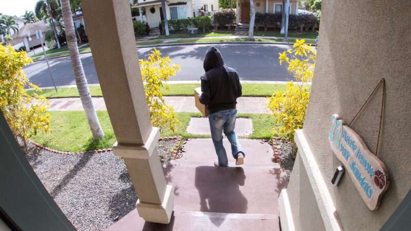 A package thief in a hoodie walks away from a porch with a package in tow, as seen through a doorbell camera.