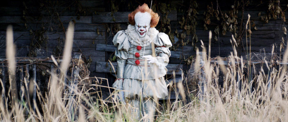 Pennywise the Dancing Clown from the movie "It" stands menacingly in front of an old building, dressed in a Victorian-style clown outfit with pom-poms