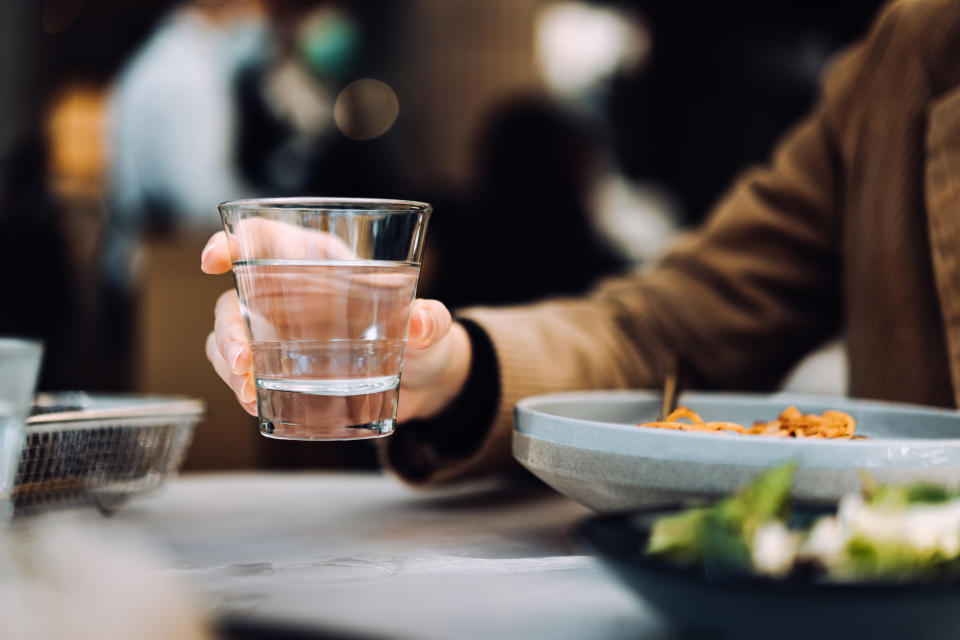 Person holding a glass of water at a restaurant table with food and blurred background