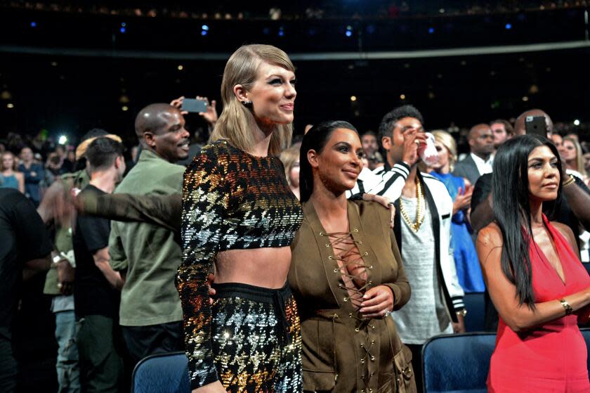 Taylor Swift, in multicolor matching set, and Kim Kardashian, in green dress, stand arm in arm in crowd