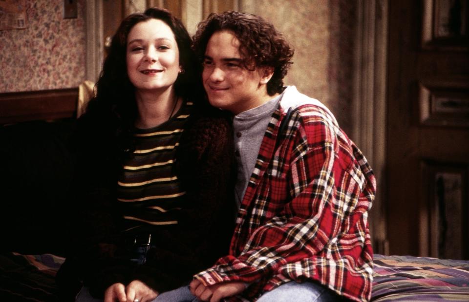 Sara Gilbert and Johnny Galecki are sitting closely on a bed, smiling. Both are in casual attire: Sara in a striped shirt and Johnny in a plaid shirt over a gray turtleneck