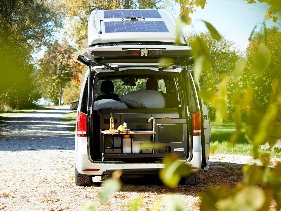 The trunk of the EQV open revealing a mini kitchen. The van is parked among trees.