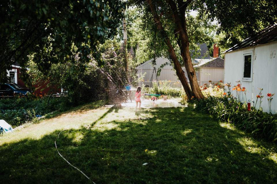 A young child plays in a backyard sprinkler on a sunny day, surrounded by trees and greenery