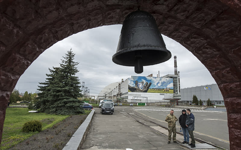 A bell is seen in the foreground as part of the monument to victims of the Chernobyl nuclear disaster, while the power plant and attendees are seen in the background