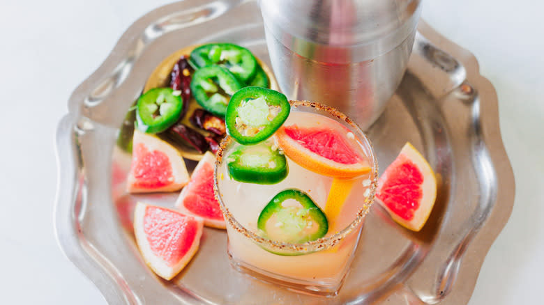 A glass of margarita with garnishes and shaker on metal tray