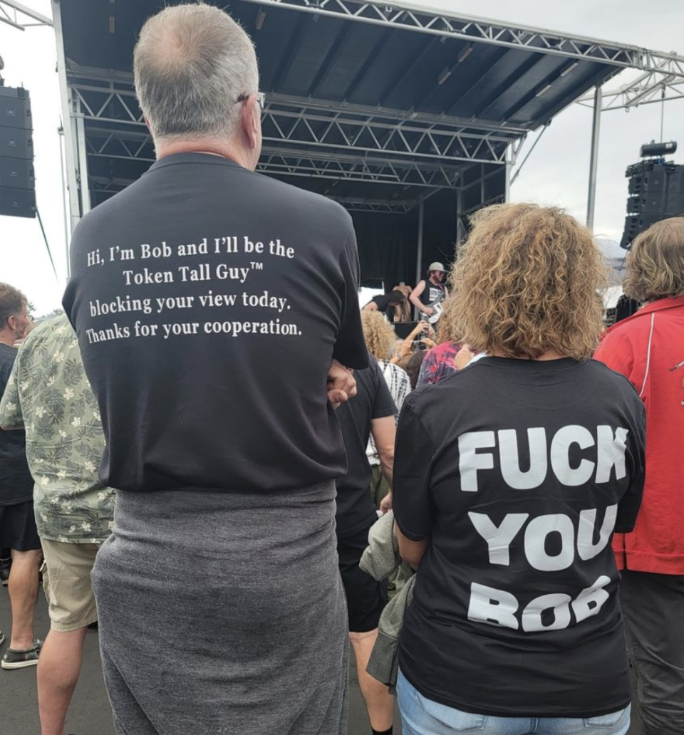 Man with "Hi, I'm Bob and I'll be Token Tall Guy blocking your view today, thanks for your cooperation" and a woman with "Fuck you Bob" on the backs of their T-shirts