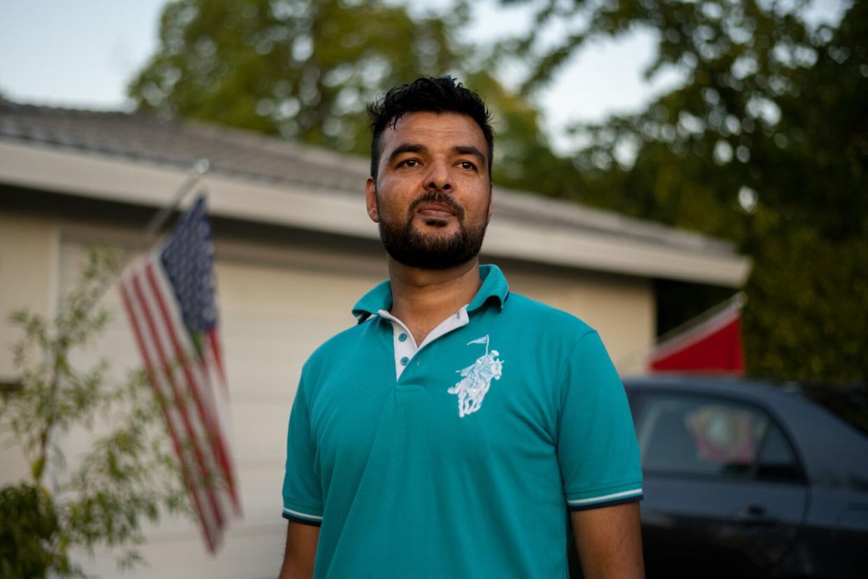 A man with dark hair and beard, in a blue polo shirt, stands outside a house flying a U.S. flag