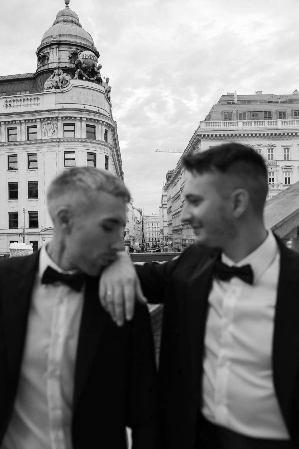 A black and white photo of two grooms looking at each other. One kisses the other's hand.