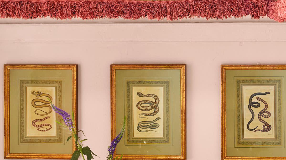 a bar covered in a patterned cloth with a plum colored background has a tray with bottles, a bowl of lemons and limes, and a small lamp, above are three framed snake prints and a shelf with colored glasses