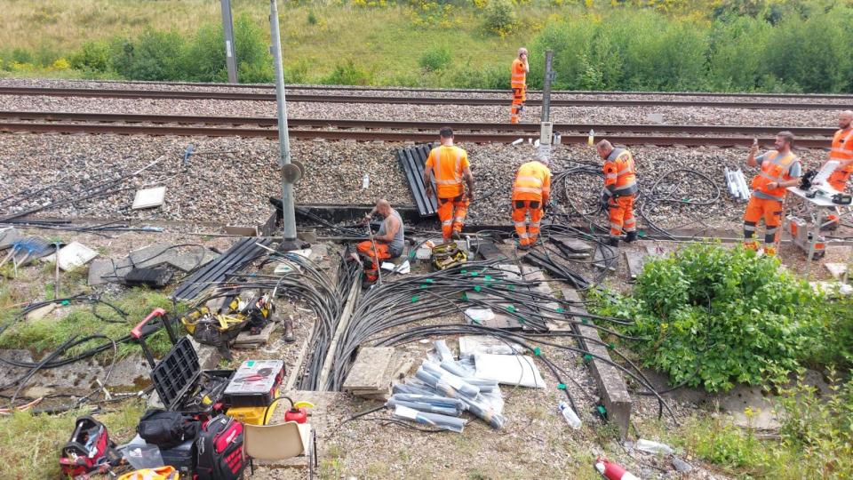 Railway workers in orange stand around the side of a rail track working to fix black cables spread over the ground.