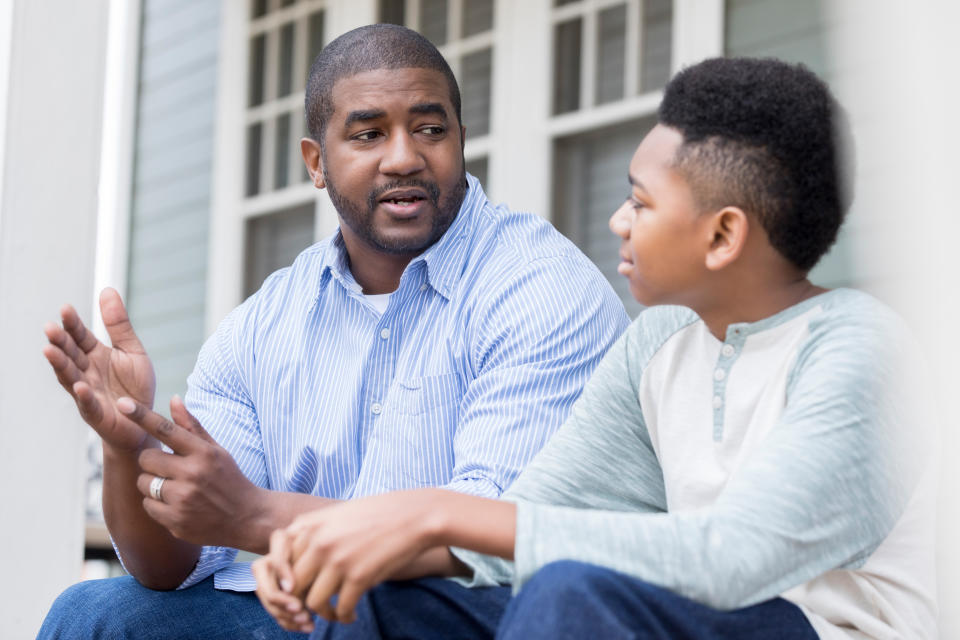 A Black man, wearing a blue and white striped shirt, sits and talks kindly to his young son, wearing a white and green T-shirt