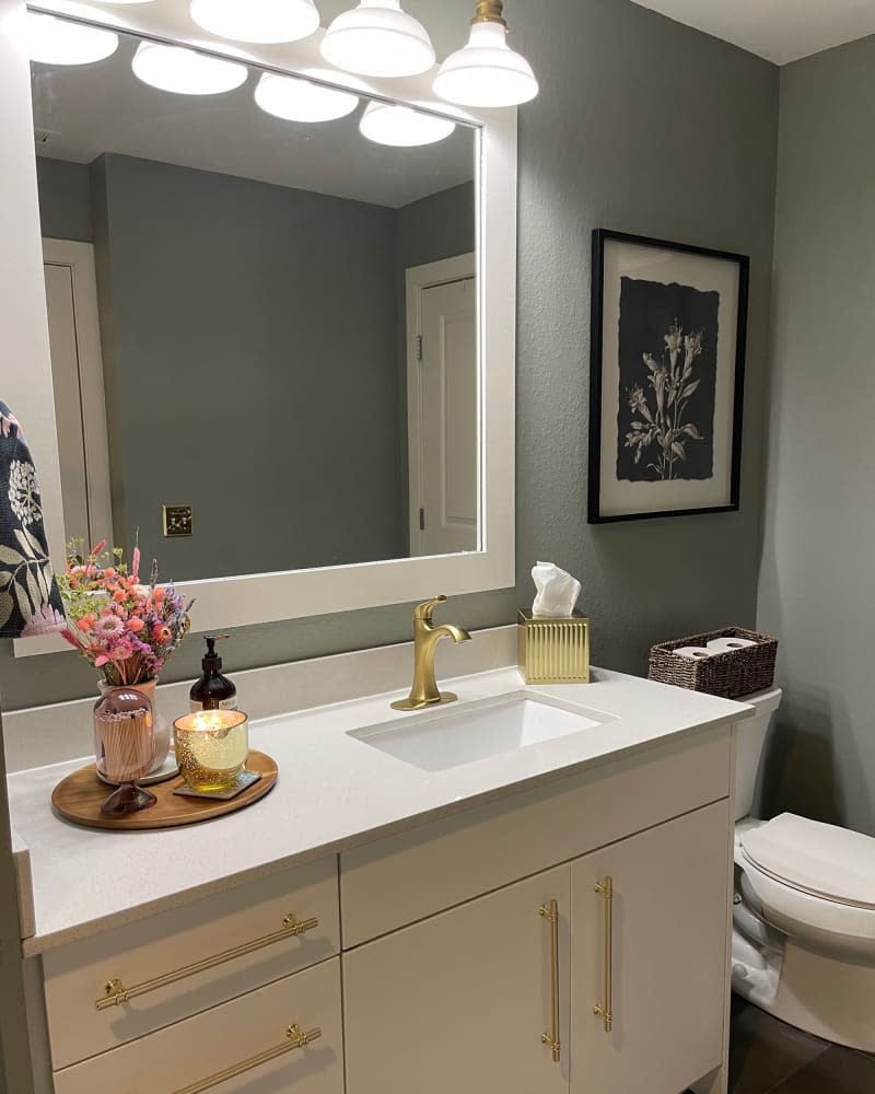 Round wood tray holding vase of flowers, matches, candle, and soap atop white sink with gold faucet and white cabinetry under white framed mirror in sage bathroom.
