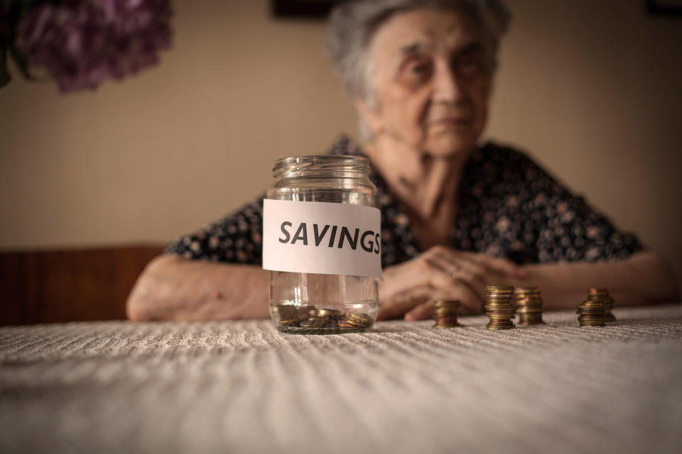 Elderly woman with near-empty jar marked "SAVINGS" sitting in front of her on the table