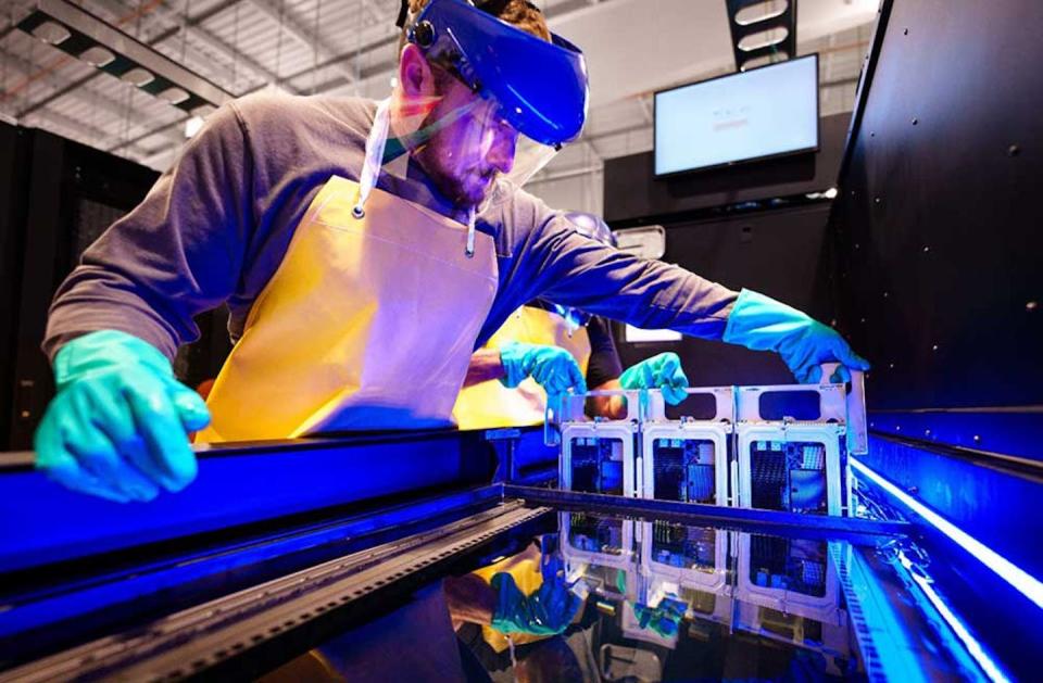 a man wearing rubber gloves and a visor lowers a circuit board into a trough containing a liquid