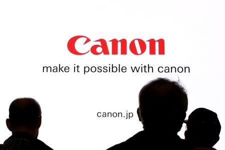 People are silhouetted against a display of the Canon brand logo at the CP+ camera and photo trade fair in Yokohama, Japan, February 25, 2016. REUTERS/Thomas Peter/File Photo