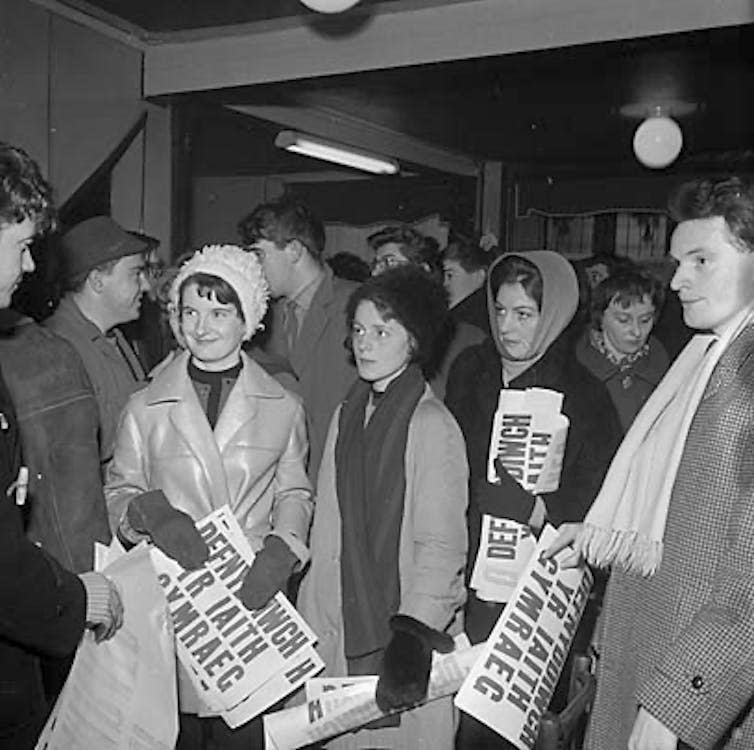 Black and white image of people in scarves, hats and coats carrying Welsh language signs saying 'defnyddiwch yr iaith Gymraeg'