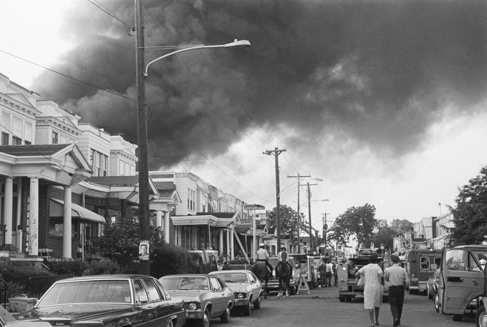 Smoke billows over rowhouses in West Philadelphia on May 12, 1985 after the police bombed a residence home to members of the Black liberation group MOVE. Police on horseback and emergency vehicles block off a street as residents walk towards the scene.<span class="copyright">Bettmann Archive—Getty Images</span>