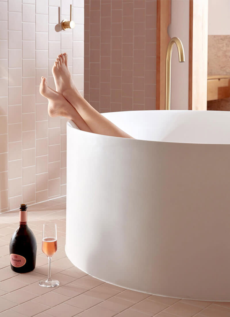 A woman's legs rest on the rim of a round tub in a bathroom, with a bottle of pink champagne and a glass on the ground. 