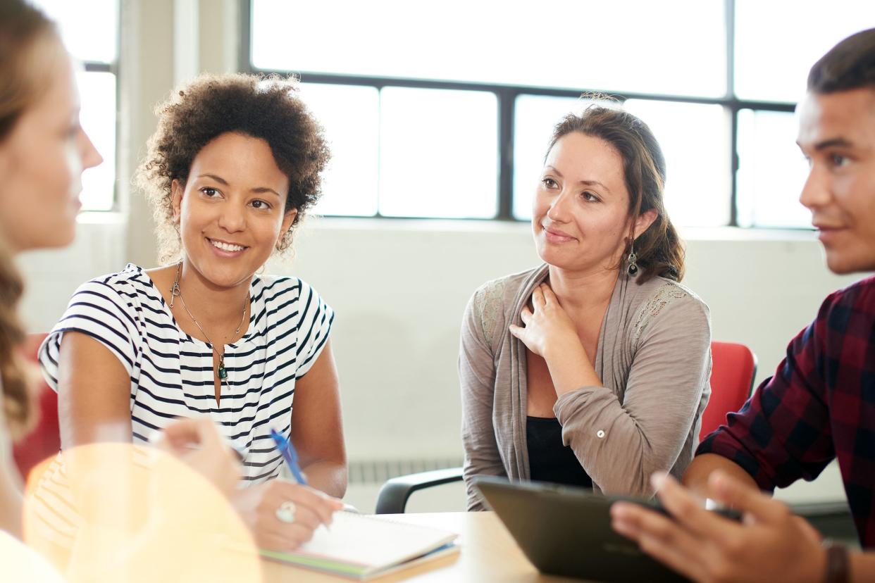 Team of four young adults sitting around a table engaging in a discussion, sitting at a desk, a man is on a tablet, a woman is writing, a large ceiling with windows blurred in the background